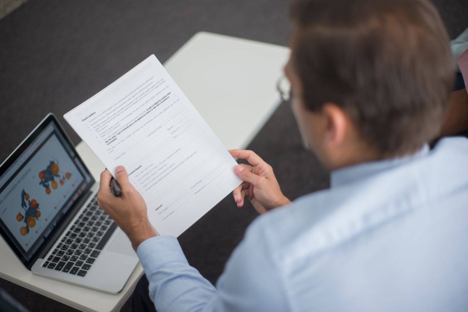 A man in a light blue shirt reads a printed form with a pen in hand, a laptop open on the table beside him.